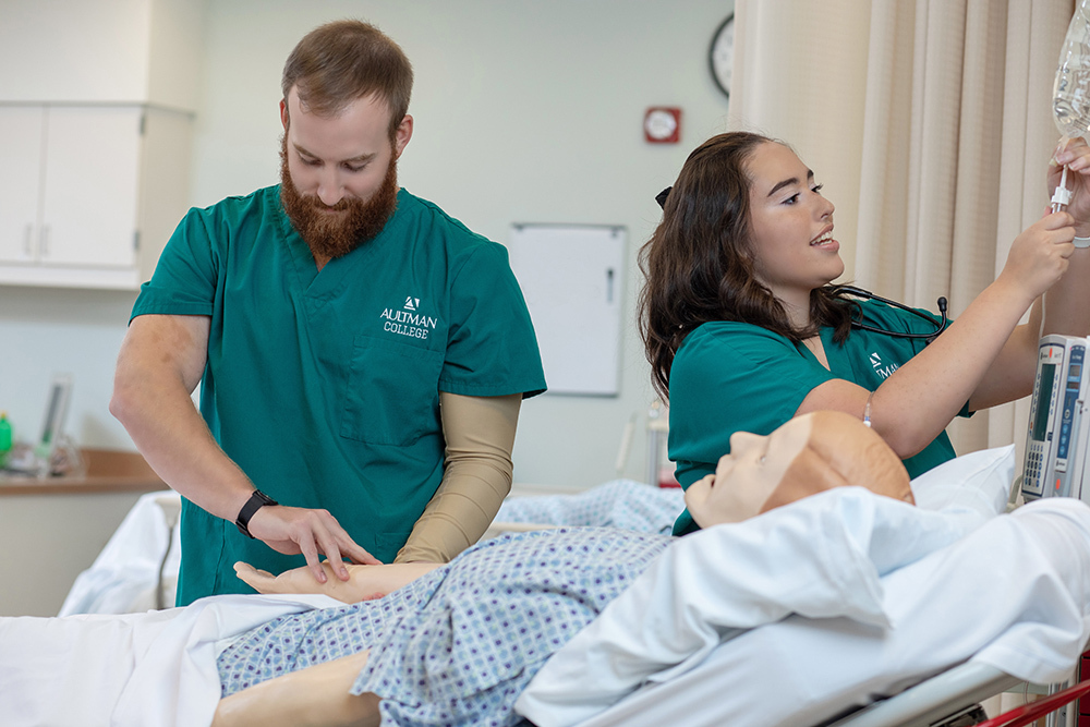 Two nursing program students train on a mannequin in a hospital bed