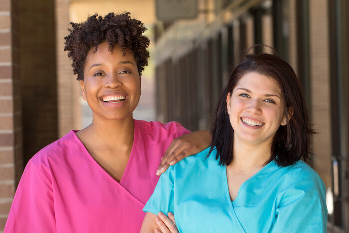 Two female nurses smiling, standing outside a healthcare clinic  